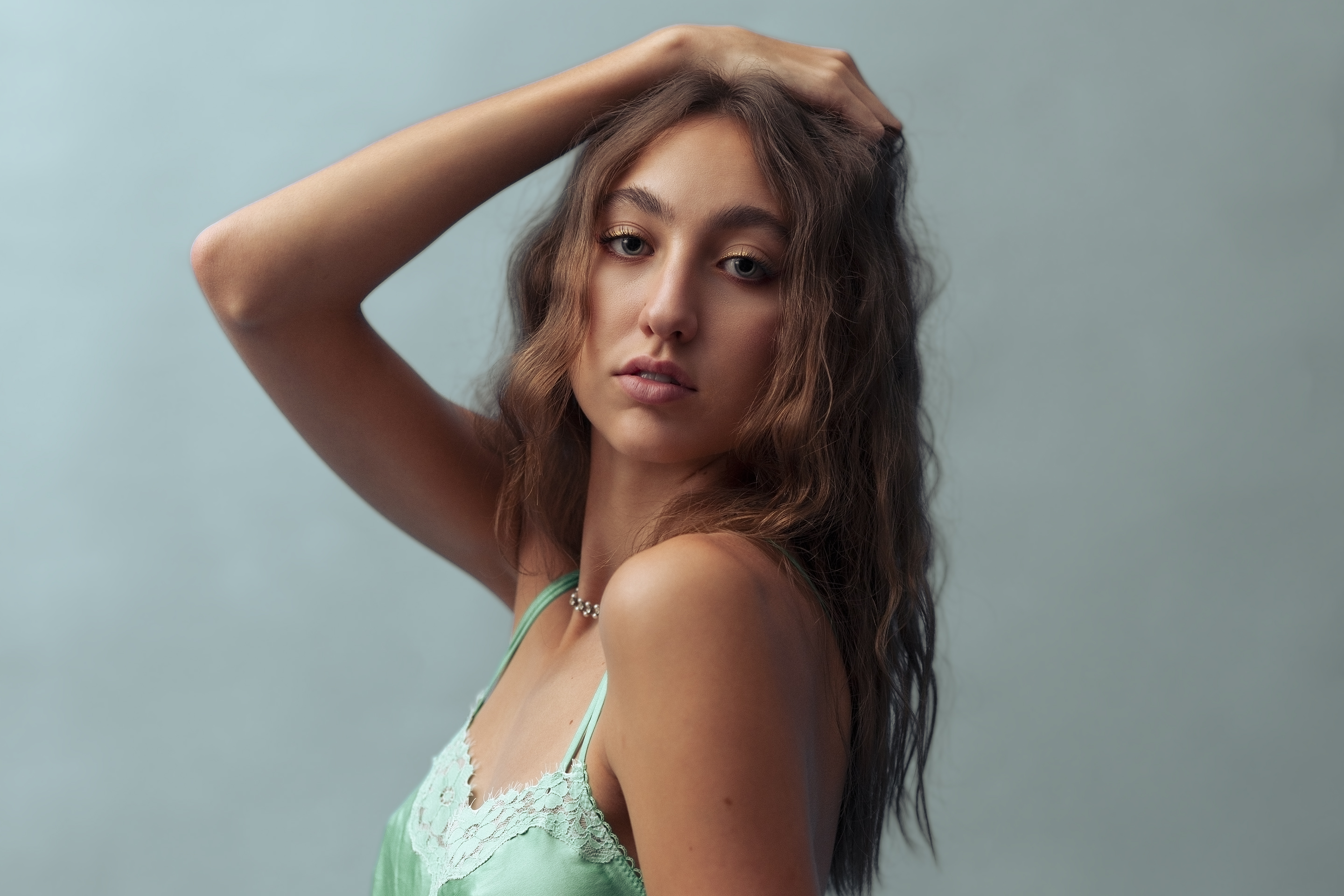 A woman with long, wavy brown hair poses with one hand on her head, wearing a light green camisole, against a plain light blue background.