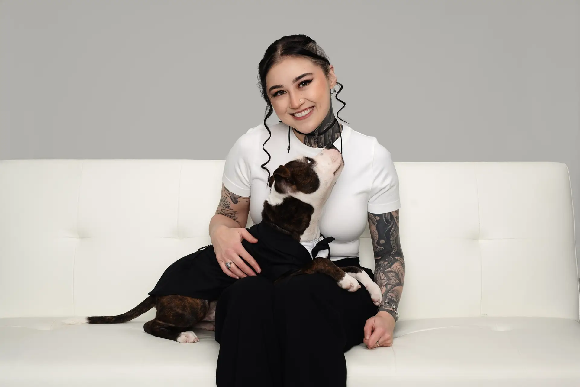 A woman with tattoos sits on a white couch, smiling at the camera, while a dog wearing a black outfit sits on her lap and looks up at her.